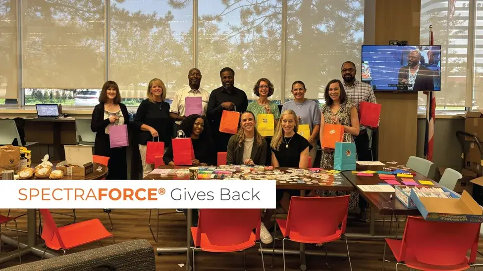 A group of employees holds colorful gift bags in a bright office setting, with decorated tables and baked goods, symbolizing the charitable efforts of SPECTRAFORCE in support of Grace Centers of Hope.