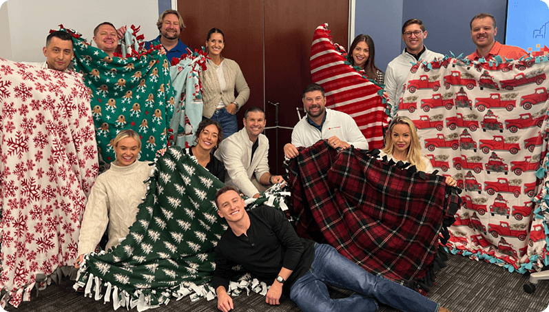 Team members in cozy sweaters holding festive blankets with holiday patterns, symbolizing warmth, community, and holiday cheer in a social responsibility context.