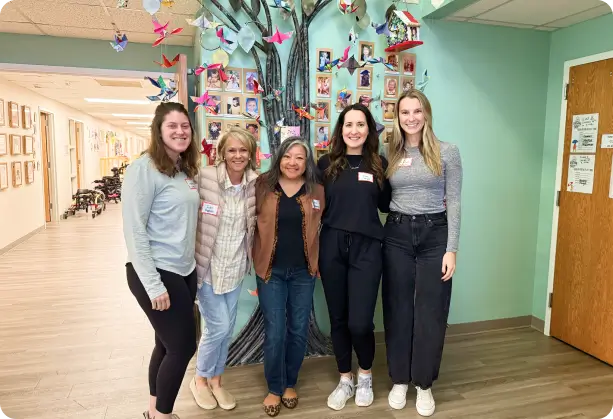 Five women standing together in front of a colorful mural of a tree decorated with photos and origami, symbolizing unity, creativity, and community in a positive work environment.