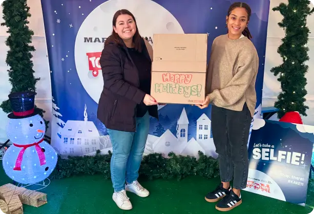 Two individuals holding a "Happy Holidays!" donation box in front of a festive backdrop with decorations and a snowman, symbolizing SPECTRAFORCE's holiday community outreach and giving.
