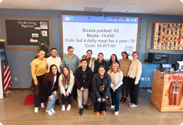 A group of individuals standing in a community center, with a presentation showcasing meal packing statistics in the background, highlighting SPECTRAFORCE's commitment to community support.