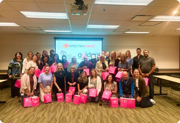 Group photo of individuals holding pink bags, with a backdrop featuring the SPECTRAFORCE and Guidant logos in a conference room, symbolizing collaboration and partnership.