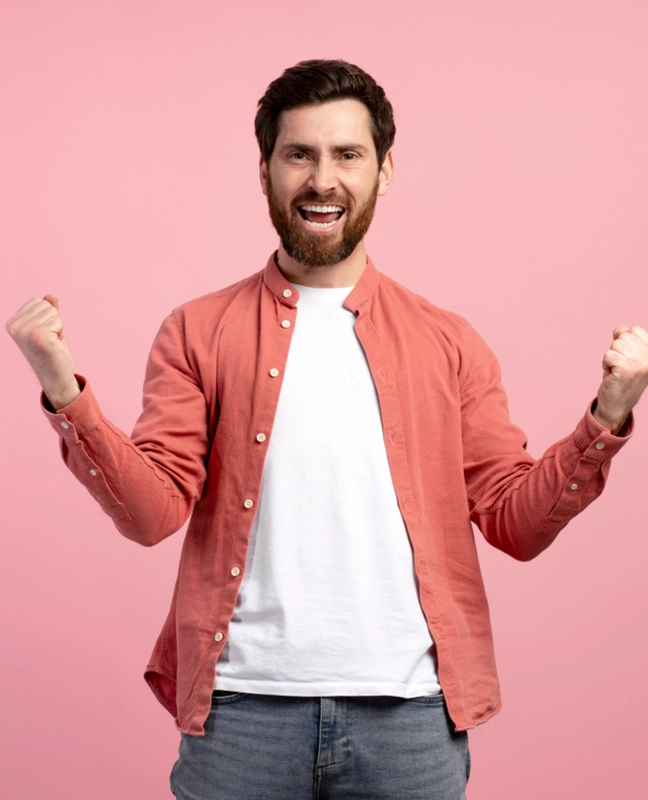 A person in a coral button-up shirt and white t-shirt raises their fists in a triumphant pose against a pink background.