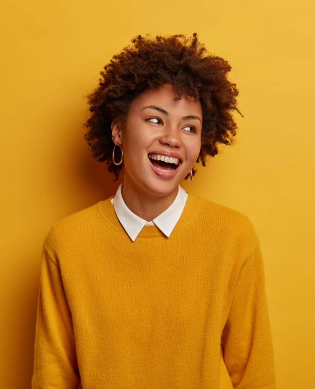 A person with curly hair wearing a yellow sweater and a white collared shirt stands in front of a vibrant yellow background.