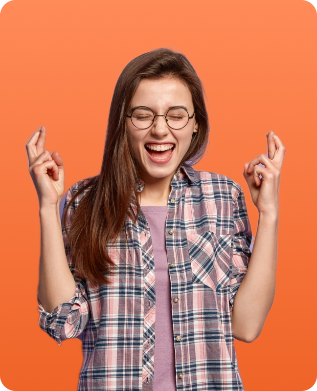A young woman wearing a plaid shirt and a purple top, holding her fingers crossed against a bright orange background.